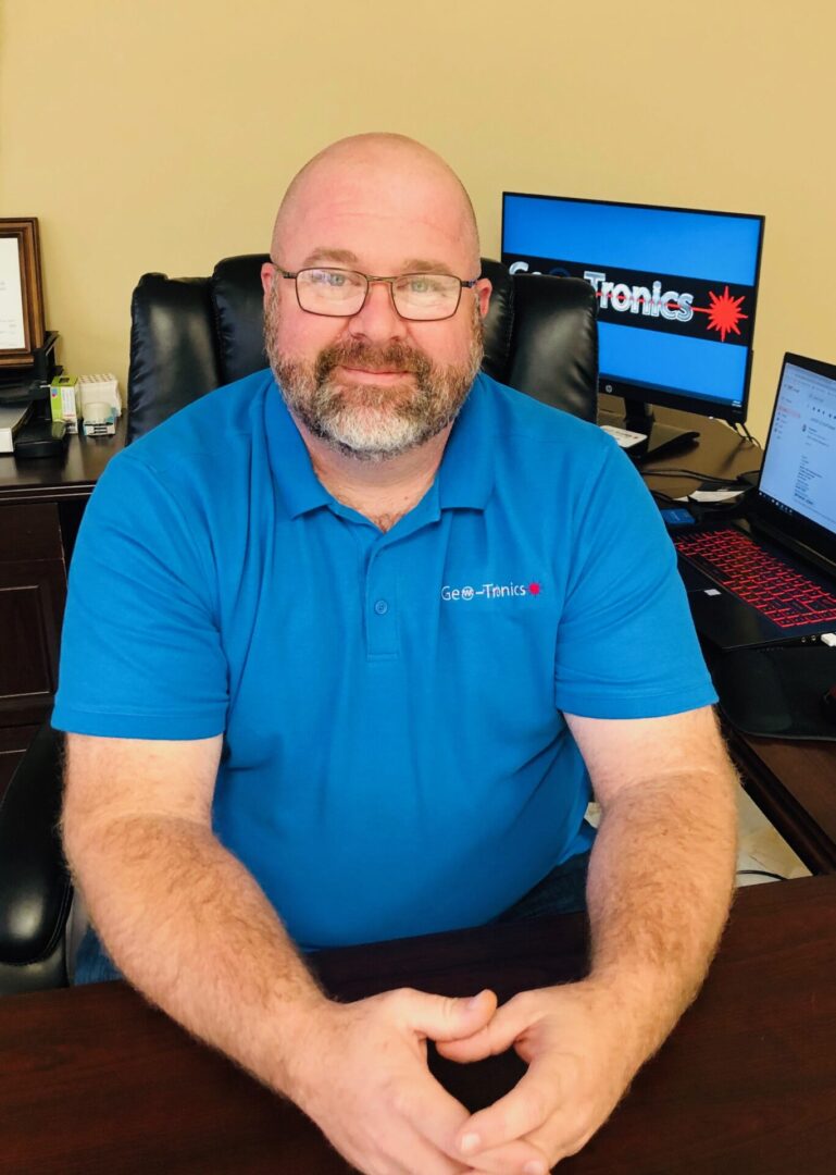 A bald man wearing a blue Geo-Tronics shirt and eyeglasses while sitting on a black, leather office chair